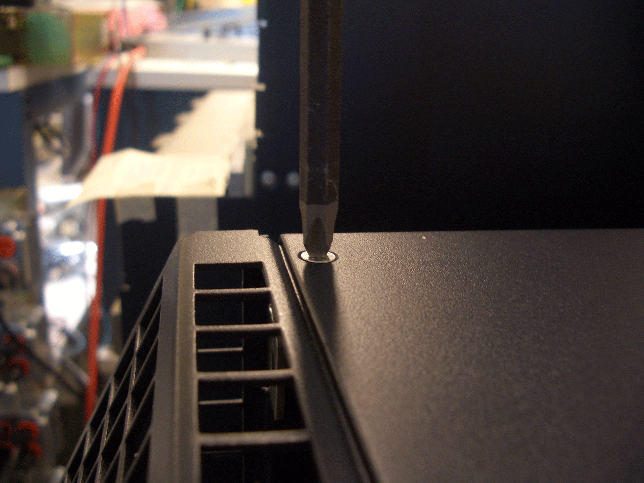 Screwdriver adjusting a screw on a black metal surface with ventilation slots, likely a computer case in a workshop.