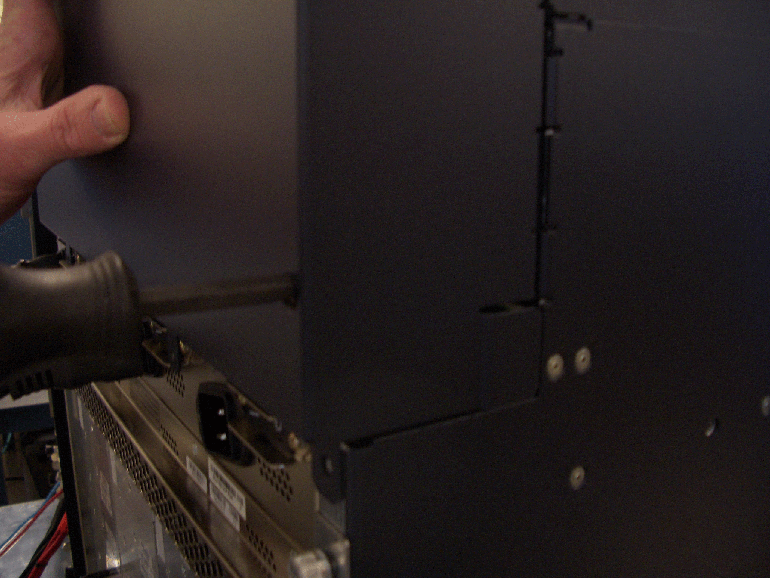 Person using a screwdriver on electronic equipment, holding a side panel with visible screws in a repair setting.